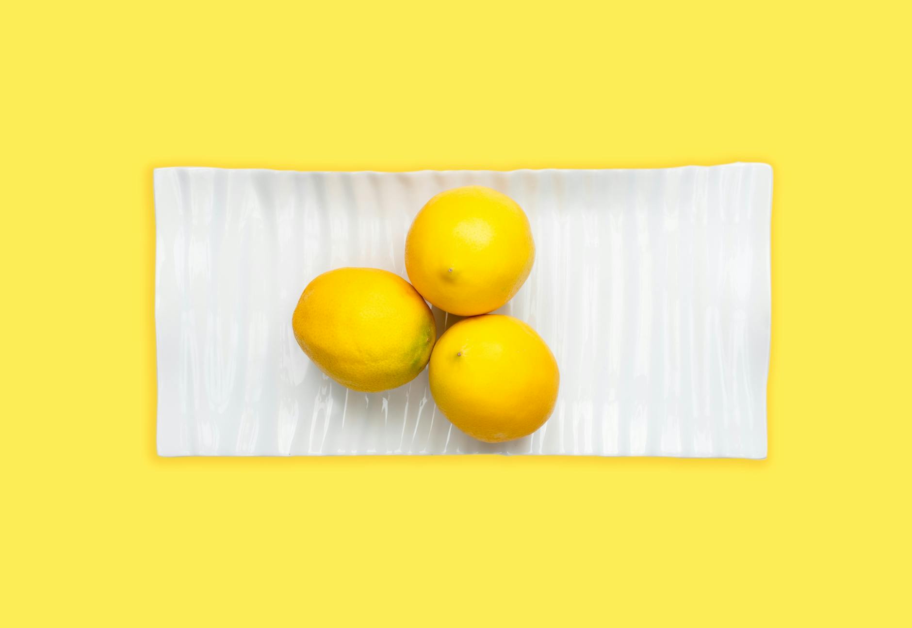 Tre limoni freschi disposti su un piatto bianco con uno sfondo giallo vivace, enfatizzando freschezza